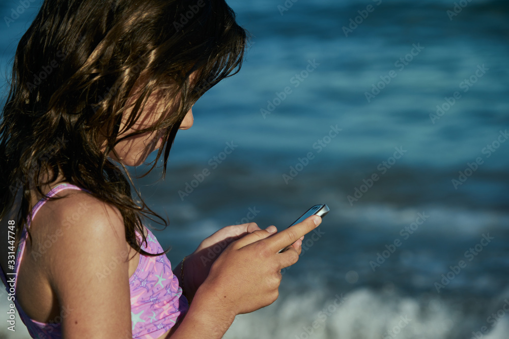 Back view of a teenage girl holding a cell phone while sitting alone on the beach enjoying the seascape on a summer day, a girl watching and writing on her cell phone