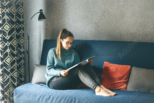 Obraz na plátne A girl reads an entertainment or educational magazine while sitting on a sofa in a cozy home environment