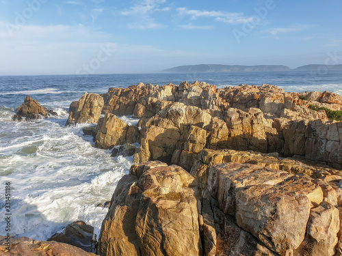 Sunlight rocks in sea foam with sea, sky and landscape in the background.