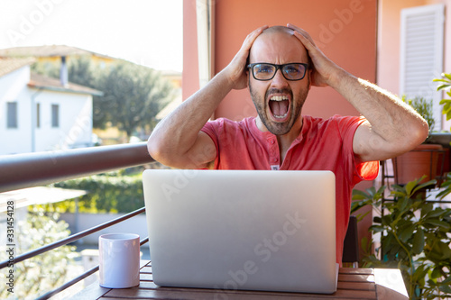 Young boy studying and working on a project outdoor on his laptop is going crazy. Very busy caucasian student sitting outside, on a wooden desk, with hands on his head. Business concept. 