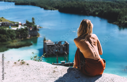 At a picturesque vantage point of Rummu quarry, a blond woman is standing in her bare feet, wearing a thigh high, auburn coloured dress, With her back turned away from the camera. Clear Water. photo