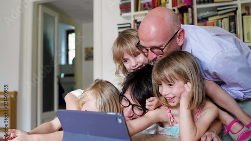 Three little sisters indoor at home using tablet sitting table supervised by parents - education, security, streaming concept