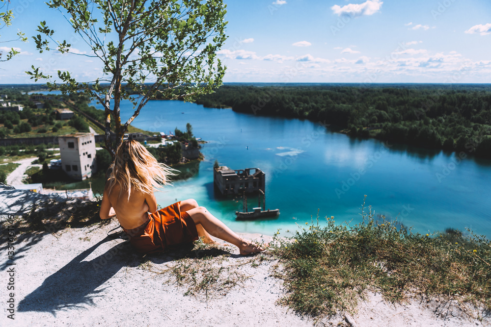 At a picturesque vantage point of Rummu quarry, a blond woman is sitting, wearing a thigh high, auburn coloured dress, With her back turned away from the camera. Clear Water.