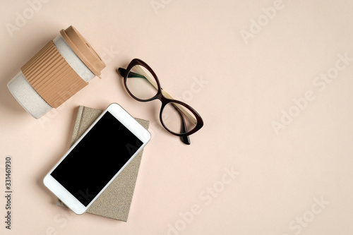 Flat lay feminine home office workspace with paper notepad, glasses, reusable bamboo coffee cup, phone with blank screen mockup. Cozy female office desk concept.