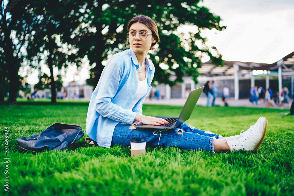 Fototapeta premium Serious caucasian female student sitting on college campus doing homework online via 4G connection, millennial young woman using laptop computer for publication in blog typing articles on rest in park