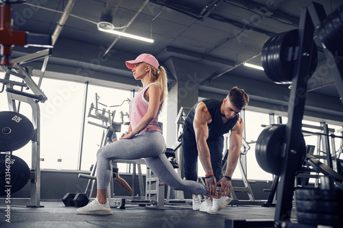 Side view of focused and motivated sporty young blonde girl in sportswear doing legs exercises while handsome muscular personal trainer monitoring her in the gym.