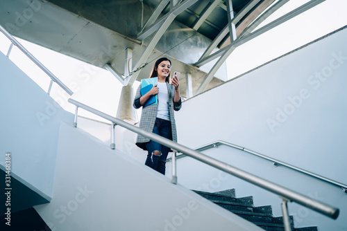 Smiling young woman reading notification on mobile phone walking down on urban staircase, cheerful trendy dressed millennial hipster girl happy about getting good 4G internet send text message
