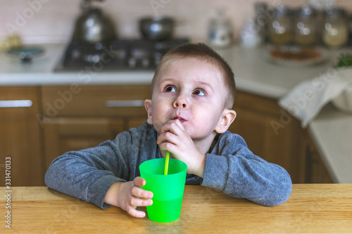 Portrait of thoughtful little boy in the kitchen at home. Pretty child drinks a drink through a plastic straw and looking up thoughtfully.