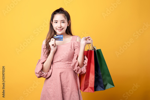 Beautiful Asian woman in red line dress are holding up a credit card and shopping bag happily in yellow seamless background. Online shopping, payment convenience, happy lifestyle concept.