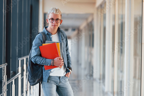 Male young student in jeans clothes is in corridor of a college with notepad in hands