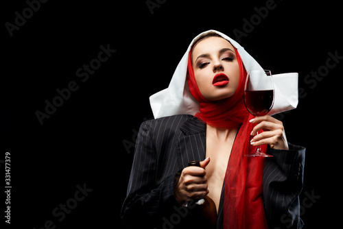 attractive sensual nun holding glass and bottle of red wine isolated on black photo