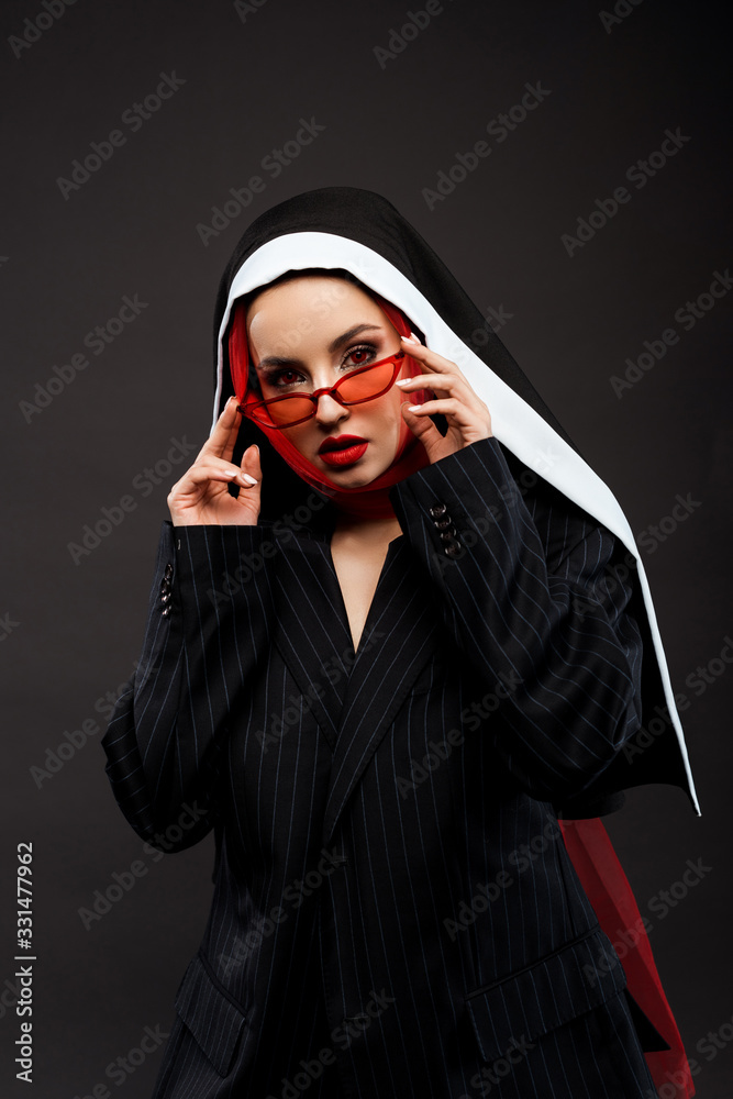 fashionable sexy demoniacal nun with red eyes posing in black suit, red sunglasses and scarf, isolated on grey fashionable sexy demoniacal nun with red eyes posing in black suit, red sunglasses and scarf, isolated on grey