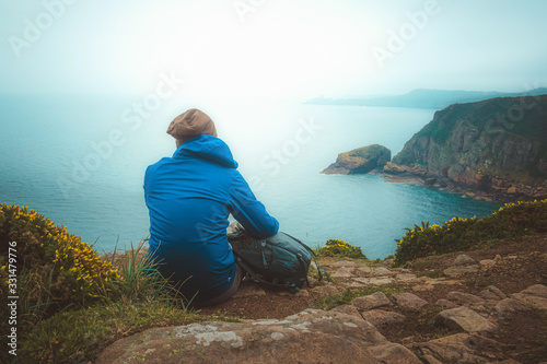 Young hiker man sitting with his backpack on the edge of a cliff to contemplate the scenic coastline landscape. Travel, lifestyle, outdoors, nature concepts.
