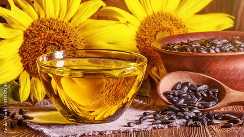 Rural still-life - the sunflower oil in glass bowl with flowers of sunflower (Helianthus annuus) and raw seeds against the background of an wooden wall, closeup