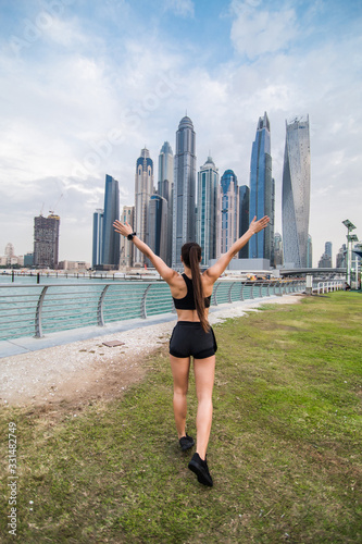 Rear view of young fitness woman with raised hands look at skyscrapers. Sport success concept.