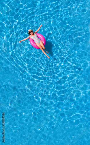 Zenith aerial view of a swimming pool in summer. Young girl in a swimsuit and hat floating with pink donut.