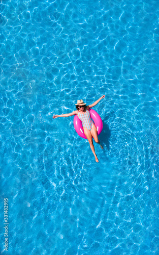 Zenith aerial view of a swimming pool in summer. Young girl in a swimsuit and hat floating with pink donut.