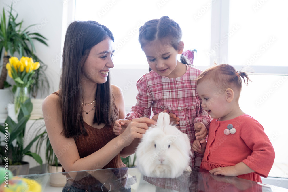 Cheerful smiley mother and her daughters playing with Easter white rabbit and laughing. Happy family preparing for Easter. Easter family concept. White window background. Yellow flowers. Cheerful smiley mother and her daughters playing with Easter white rabbit and laughing. Happy family preparing for Easter. Easter family concept. White window background. Yellow flowers.