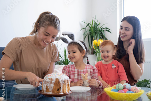 Two beautiful mothers with their daughters celebrating Easter eating cake and having fun together on a sunny morning. Girls having breakfast in a white dining room with a big window. Easter concept.
