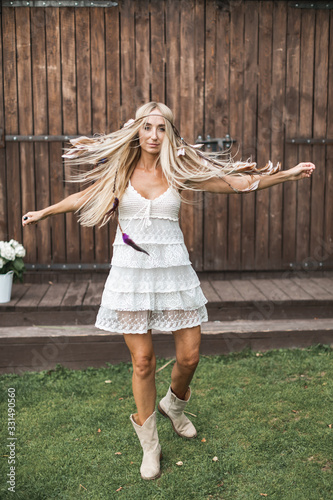 Pretty young boho-style blond woman, wearing feathers in the hair, white dress and cowboy boots, standing happy with open arms, in front of stylish vintage wooden barn