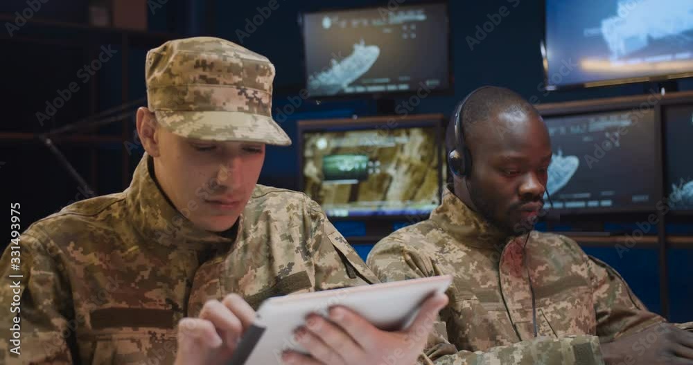 African American young man in headset typing on laptop computer and Caucasian soldier tapping on tablet device in control room. Multiethnic army officers in uniforms working together in office.