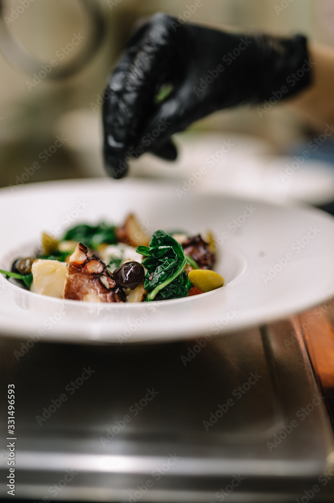 Final touches in the preparation of the salad. The chef adds some spices to octopus salad with stracciatella, tomatoes, olives and green basilisk on a plate. Smooth image with shallow depth of field.