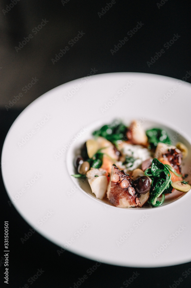 Octopus salad with stracciatella, tomatoes, olives and green basilisk on a plate. Dark table background. Smooth image with shallow depth of field.