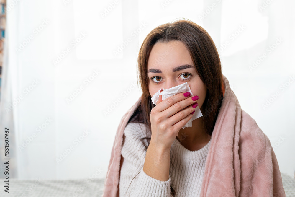 A young Caucasian woman, covered with a blanket, covers her face with a handkerchief. Treatment at home. Copy space. Concept of viral infection and quarantine A young Caucasian woman, covered with a blanket, covers her face with a handkerchief. Treatment at home. Copy space. Concept of viral infection and quarantine