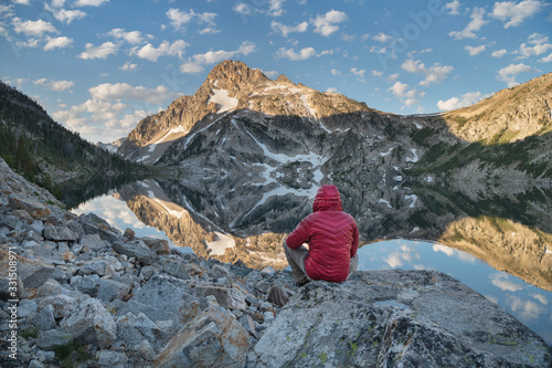 Adult male hiker in red jacket admired the view of Mount Regan mirrored in still waters of Sawtooth Lake at sunrise. Sawtooth Mountains Wilderness Idaho; Adult male hiker in red jacket at Sawtooth Lake. Sawtooth Wilderness Idaho. Idaho. United States