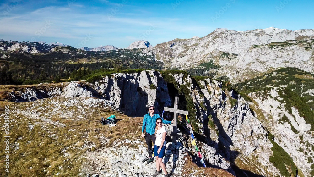 A drone shot of a couple in hiking outfits standing next to a cross on top of Buchbergkogel, Hochschwab region, Austria. They are surrounded by mountains. Achieving a goal. Alpine landscape.