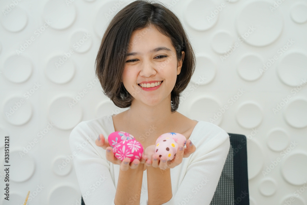 portrait of young attractive joyful smiling Asian Thai korean woman painted Easter colorful egg and sitting on the table with white background portrait of young attractive joyful smiling Asian Thai korean woman painted Easter colorful egg and sitting on the table with white background