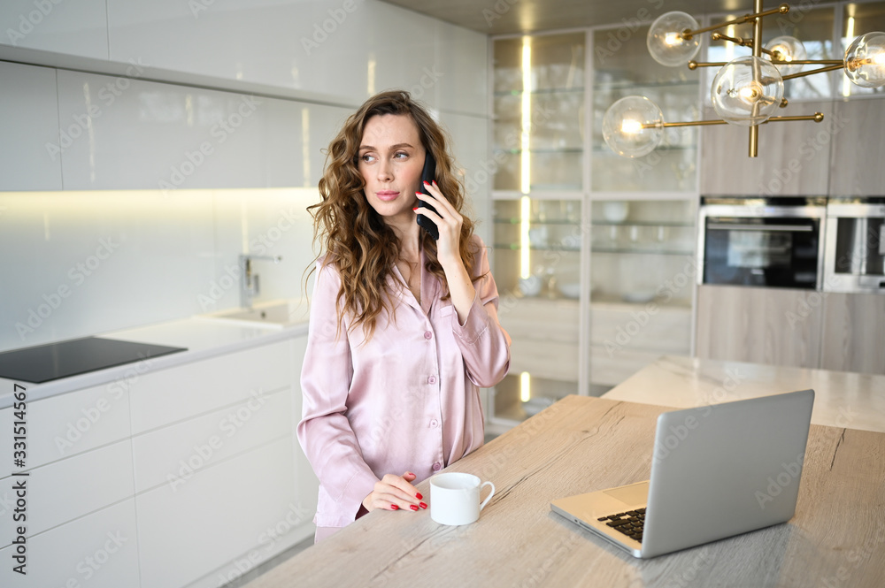 custom made wallpaper toronto digitalYoung woman speaking smartphone and working on a laptop while drinking morning coffee. Home quarantine during Covid-19 pandemic Corona virus. Distance work from home concept. Modern kitchen interior