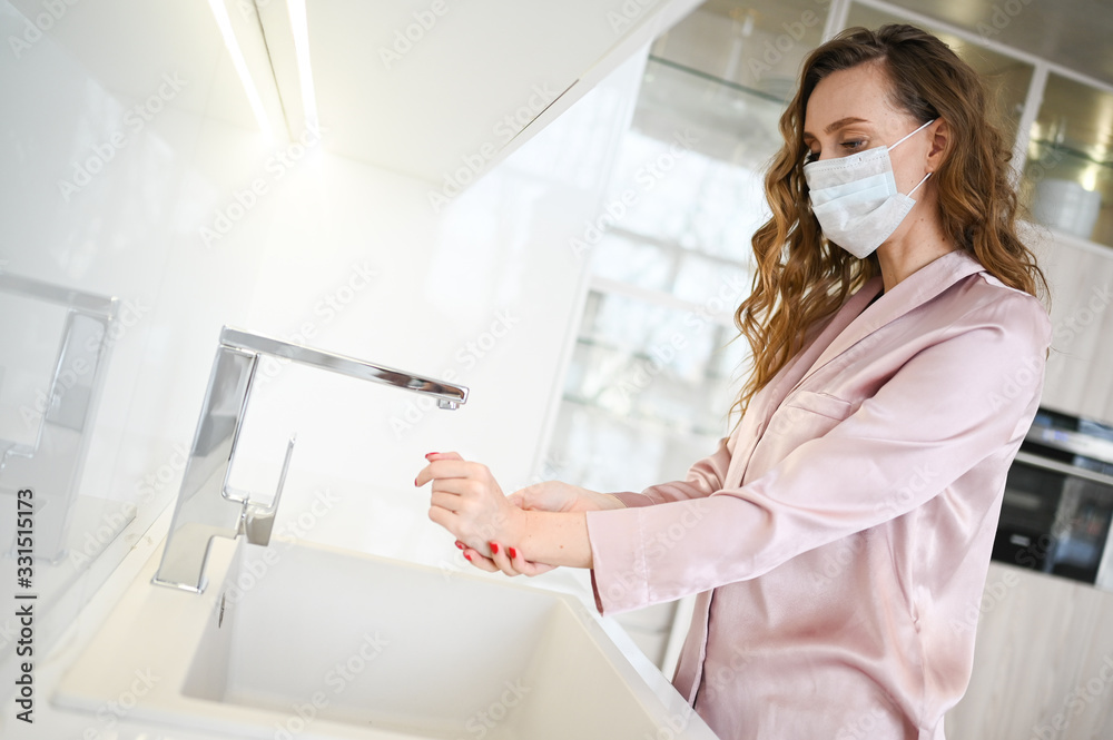 Young woman in face mask washing hands for corona virus prevention, hygiene to stop spreading coronavirus. Avoid contaminating Corona virus Covid-19 being in home quarantine in white kitchen interior Young woman in face mask washing hands for corona virus prevention, hygiene to stop spreading coronavirus. Avoid contaminating Corona virus Covid-19 being in home quarantine in white kitchen interior