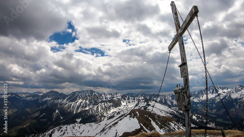 Wooden cross at the top of Himmeleck in Austrian Alps. There is a massive mountain range in the back, partially covered with snow. Early spring vibes. barren mountain slopes. Overcast. Achievement © Chris