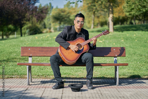 Hispanic male musician playing acoustic guitar sitting on a park bench with his black hat on the floor