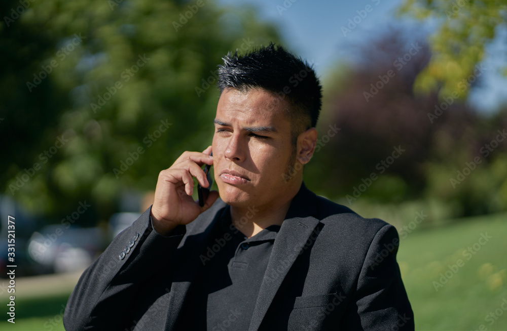 Hispanic man dressed as an executive in a black suit sitting on a park bench and talking on a cell phone Hispanic man dressed as an executive in a black suit sitting on a park bench and talking on a cell phone