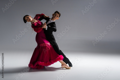 elegant young couple of ballroom dancers in red dress in suit dancing on grey