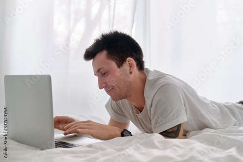 Young man in the white shirt on a bed with white linen is upset looking at the screen of a laptop.