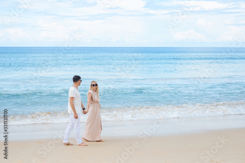 Young attractive man and woman in love walk and hug against the background of white sand and azure sea. Travel, honeymoon