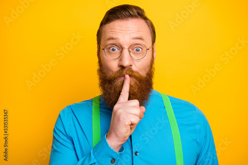 Closeup photo of funny red head guy tricky mood hold finger on lips asking not talk say speak tell wear specs bright blue shirt green suspenders isolated yellow color background photo