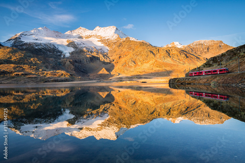  The Swiss train reflected in the white lake in the background the peaks of the Alps, Bernina Pass, Switzerland