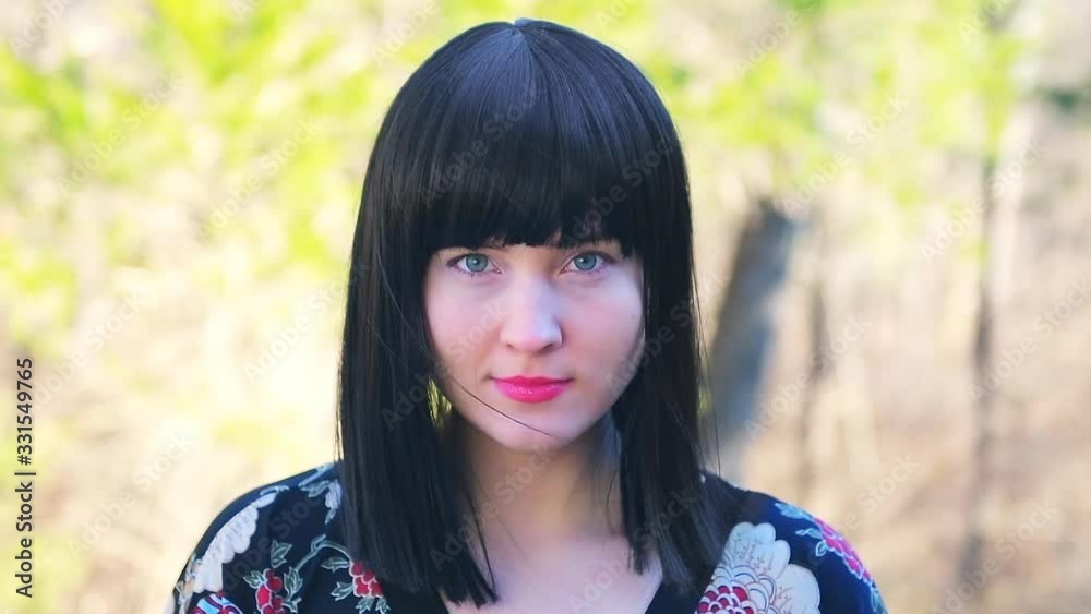 Closeup of young happy Caucasian woman in black hair Asian wig head and shoulders standing outside outdoors in outdoor park garden with colorful trees forest sunny background