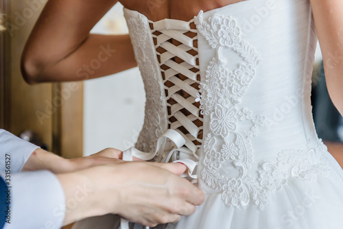 Bridesmaid or bride's mother makes bow-knot on the back of brides wedding dress. Preparation for the wedding day before the ceremony. Beautiful lace back of the white dress