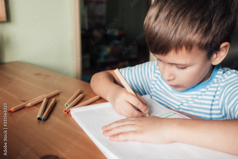 Cute boy sits at desk and draws in album with colored pencils, lifestyle