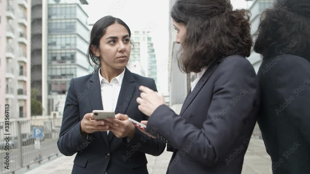 Businesswomen talking and using smartphones on street. Concentrated female colleagues using mobile phones while standing outside urban building. Technology concept Businesswomen talking and using smartphones on street. Concentrated female colleagues using mobile phones while standing outside urban building. Technology concept