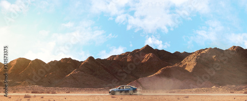 a scenery of a car riding in the center of a sandy road among the mountains with cloudy sky