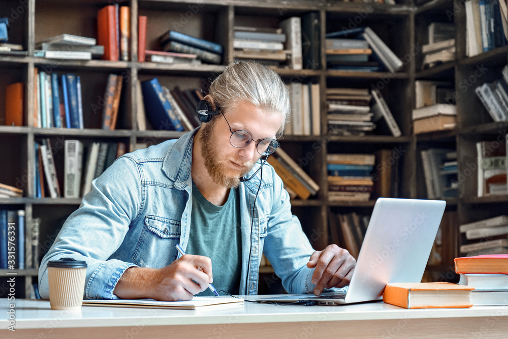 Focused young man student wear headset make notes e study online in library listen lecture internet course training online learning foreign language distance education write information in notebook. Focused young man student wear headset make notes e study online in library listen lecture internet course training online learning foreign language distance education write information in notebook.