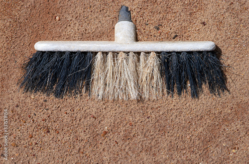 The old discarded head of a black and white plastic broom lies in red sandy dirt, recent rain texturing the surface, making it appear as if the broom is somewhat sunken or absorbed into the ground. 