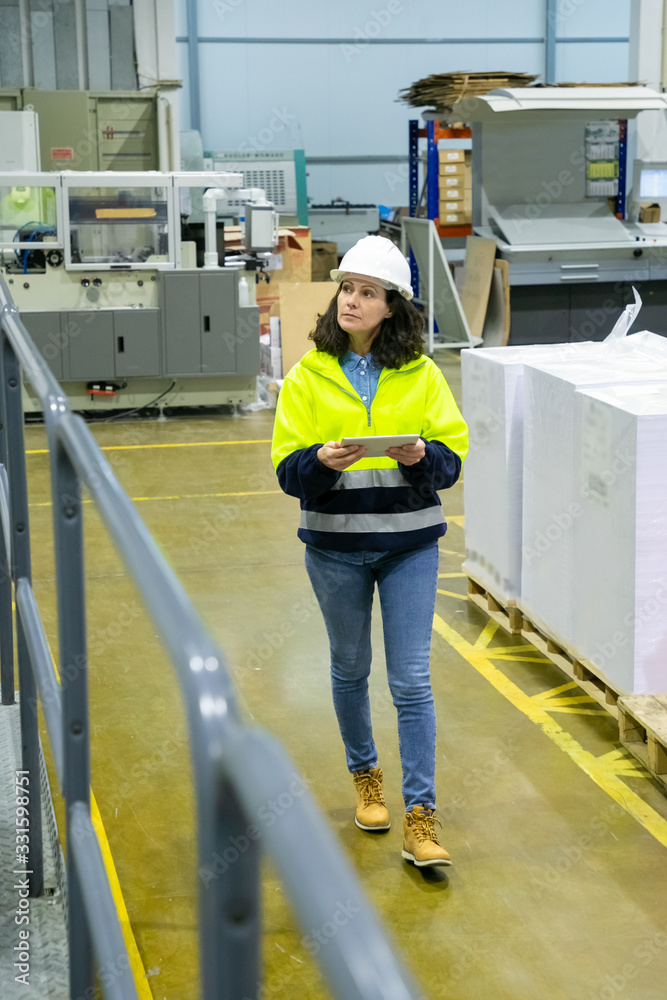 High angle view of worker using tablet while walking at factory. Focused mature woman checking equipment at printing house. Print manufacturing concept High angle view of worker using tablet while walking at factory. Focused mature woman checking equipment at printing house. Print manufacturing concept