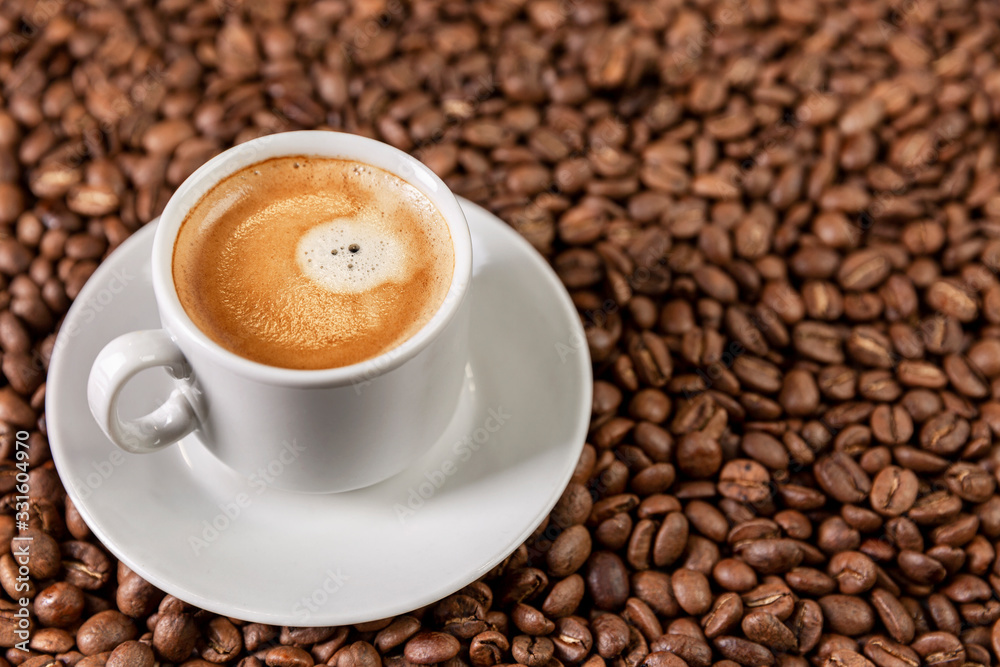 A white cup with aromatic coffee stands on scattered grains. Close-up. Space for text. A white cup with aromatic coffee stands on scattered grains. Close-up. Space for text.
